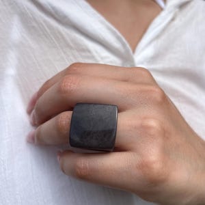 May include: A large, square, dark gray ring. The ring is worn on a finger and has a smooth, polished surface. The ring's shape is a prominent feature, and the color is a deep, neutral tone.