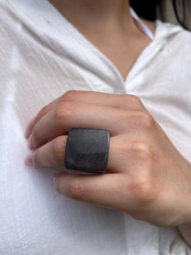 May include: A large, square, dark gray ring. The ring is worn on a finger and has a smooth, polished surface. The ring's shape is a prominent feature, and the color is a deep, neutral tone.