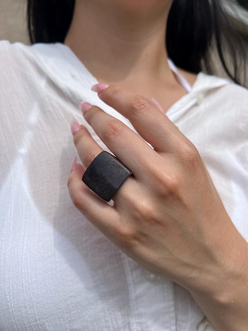 May include: A dark gray, square-faced ring. The ring is worn on a finger, and the background is a white shirt. The ring's design is simple and modern, with a flat, rectangular face.