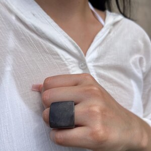 May include: A dark gray square ring. The ring is worn on a hand, and the square face of the ring is prominent. The ring is a solid color and appears to be made of a smooth material. The background is a white shirt.