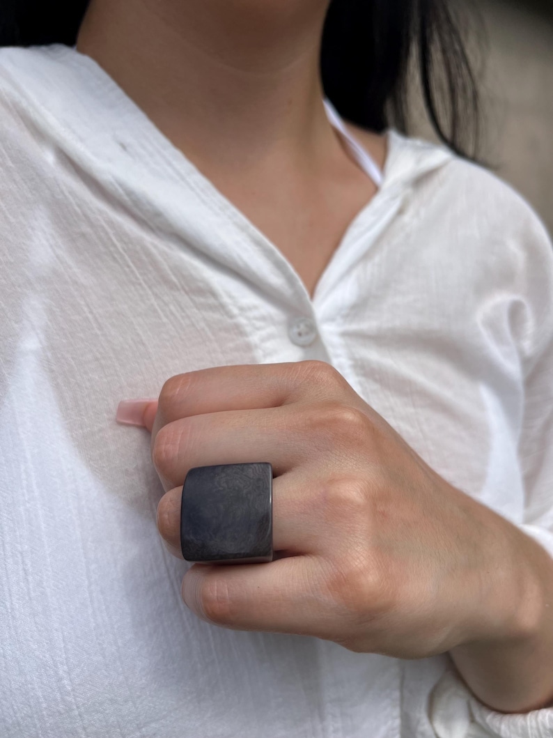 May include: A dark gray square ring. The ring is worn on a hand, and the square face of the ring is prominent. The ring is a solid color and appears to be made of a smooth material. The background is a white shirt.