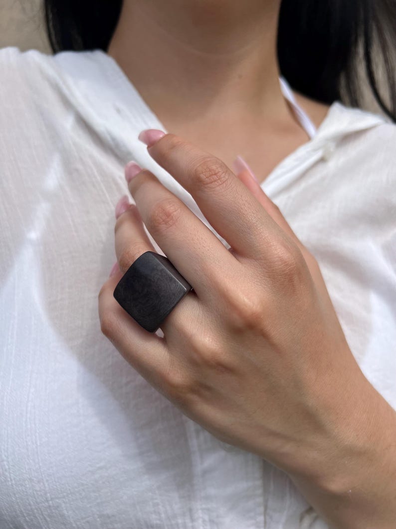 May include: A dark gray, square-shaped ring. The ring is worn on a finger. The ring is made of a solid, possibly carved material. The background is a white shirt.