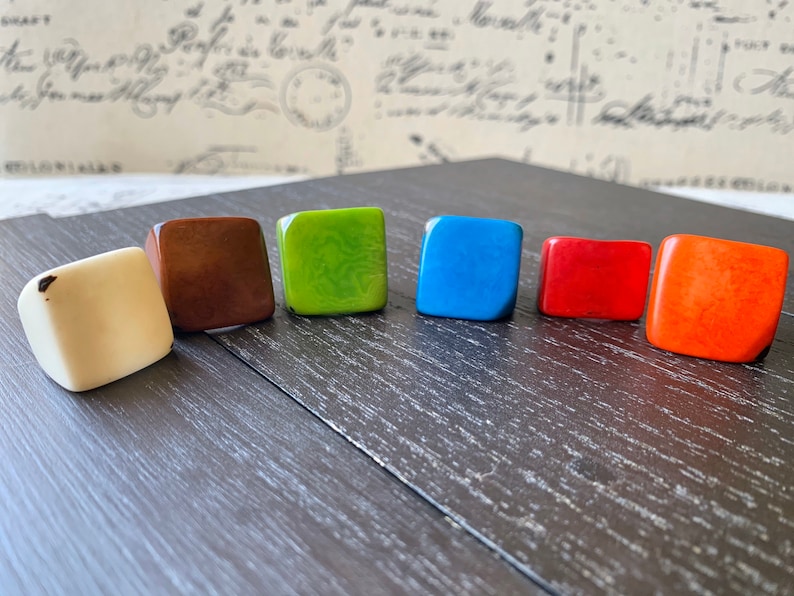 May include: Six square-shaped rings in different colors: white, brown, green, blue, red, and orange. The rings are arranged in a semi-circle on a dark wooden surface.