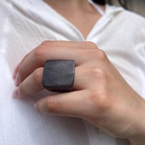 May include: A dark gray, square-shaped ring. The ring is worn on a finger. The ring has a smooth, polished surface. The ring is a statement piece of jewelry.