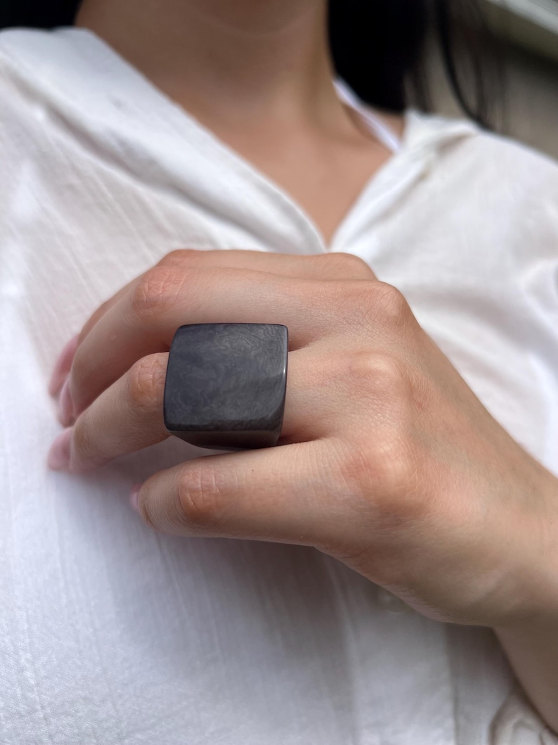 May include: A dark gray, square-shaped ring. The ring is worn on a finger. The ring has a smooth, polished surface. The ring is a statement piece of jewelry.
