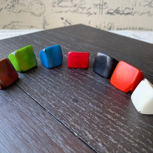 May include: A collection of colorful, square-shaped rings in various hues, including brown, green, blue, red, black, orange, and white. The rings are arranged on a dark wooden surface, showcasing their geometric design and vibrant colors.