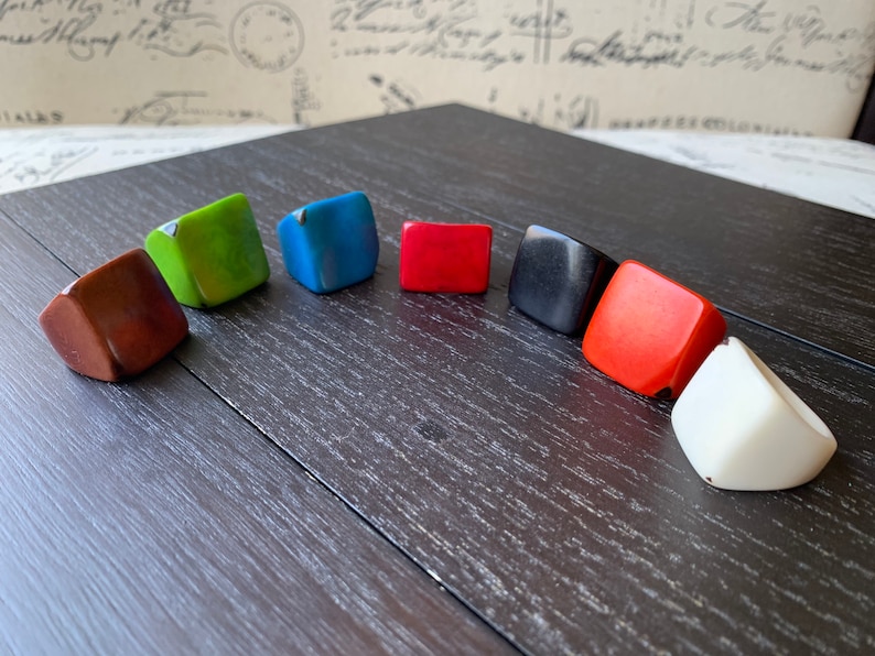 May include: A collection of colorful, square-shaped rings in various hues, including brown, green, blue, red, black, orange, and white. The rings are arranged on a dark wooden surface, showcasing their geometric design and vibrant colors.