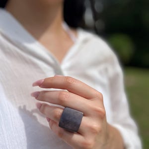 May include: A large, square, dark gray ring. The ring is worn on a finger, and the background is blurred, with a white shirt and green foliage visible.