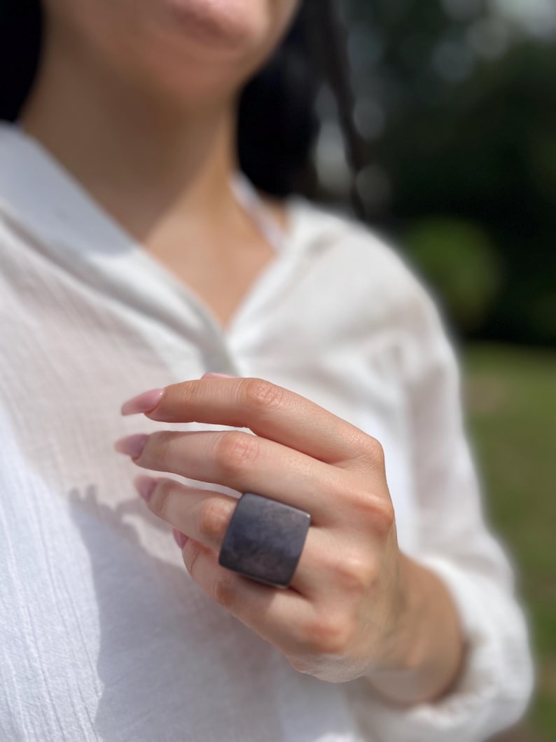 May include: A large, square, dark gray ring. The ring is worn on a finger, and the background is blurred, with a white shirt and green foliage visible.