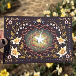 May include: A dark brown card with a white star in the center surrounded by red mushrooms and a wreath of white flowers. The card has a gold border with a repeating pattern of flowers, leaves, and dots.