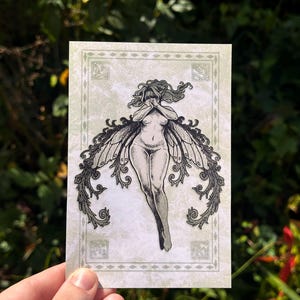 May include: A grayscale illustration of a fairy with large, ornate wings. The fairy is nude, with hands covering the face. The artwork is on a rectangular card with a decorative border and a light green background.
