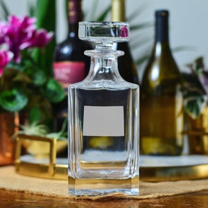 May include: A clear glass decanter with a square base and a stopper. The decanter is empty and has a label with a white rectangle in the center.