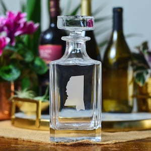 May include: A clear glass decanter with a square base and a stopper. The decanter features an etched design of the state of Mississippi.