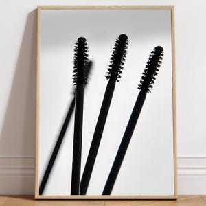 May include: A framed print featuring three black mascara wands against a white background. The wands are angled, with their bristled tips prominently displayed. The frame is a light wood color, adding a natural touch to the minimalist aesthetic.