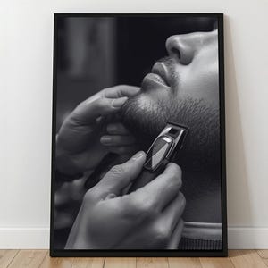 May include: Monochrome framed print depicting a barber trimming a beard with an electric trimmer. The image is a close-up of the face and hands, highlighting the grooming process. The print is displayed on a light wood floor.