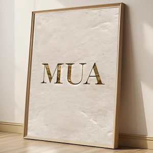 May include: A framed print featuring the letters "MUA" in a gold color, set against a textured, off-white background. The frame is a light gold color. The print is leaning against a wall.