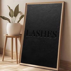Puede incluir: Un cuadro negro texturizado con la palabra "LASHES" en relieve. El marco es de color madera clara. Una pequeña maceta con una planta se encuentra sobre una mesita auxiliar de madera junto a la obra.