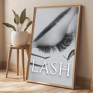 Può includere: Una stampa in bianco e nero incorniciata con un primo piano di un occhio chiuso con lunghe ciglia scure. La parola "LASH" è visualizzata in lettere bianche e in grassetto. La cornice è di colore legno chiaro e la stampa è appoggiata a un muro.