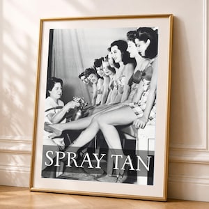 Puede incluir: Una fotografía enmarcada en blanco y negro que muestra una fila de personas recibiendo un bronceado en aerosol de un técnico. La imagen muestra el texto "SPRAY TAN" en letras mayúsculas blancas en la parte inferior. El marco es dorado.