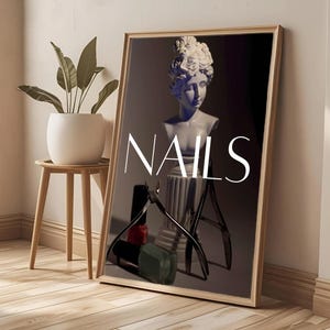 以下が含まれることがあります： 白い文字で「NAILS」と書かれた額入りのプリント。画像には白い胸像、マニキュアのボトル、爪切りが含まれています。プリントは壁に立てかけられており、木製のサイドテーブルには小さな植物が置かれています。