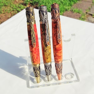 Könnte beinhalten: Drei handgefertigte Stifte mit aufwendigen Metallarbeiten und farbenfrohen, gemusterten Schäften. Die Stifte werden in einem transparenten Acrylständer ausgestellt. Die Schäfte weisen Farbtöne von Rot, Orange und Gelb auf. Die Stifte haben dekorative Metallakzente und einen Edelstein an der Spitze.