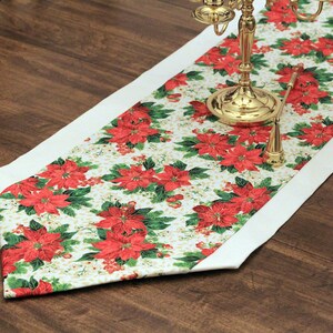May include: A white table runner with a red poinsettia print and a white border. The runner is decorated with a gold candlestick and a gold candle snuffer.