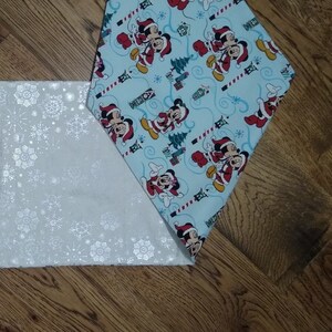 May include: A white table runner with a snowflake pattern and a blue table runner with a Christmas-themed pattern featuring Mickey Mouse and Minnie Mouse.