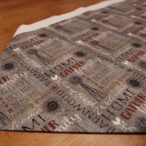 May include: A gray and white fabric table runner with a repeating pattern of text and a windmill design. The text includes the words "home", "gather", "family", "blessed", "beyond measure", "locally grown", "welcome", "barnwood", "good fresh", "faith", and "country", among others.
