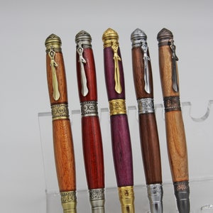 May include: Five wooden pens with metal accents and decorative tops. The pens are arranged in a row on a clear acrylic display stand. The pens are all different colors and have different designs on the tops.