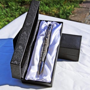 May include: A ballpoint pen with a dark, marbled gray barrel and polished silver-tone metal accents, including a clip and tip. The pen is presented inside a rectangular black box with a textured exterior and a white satin-lined interior.