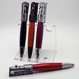May include: A selection of handcrafted pens with a train engine design. The pens have silver-coloured metal accents and wooden barrels in dark brown, orange, and purple. One pen lies horizontally, displaying its red barrel and engine detail.