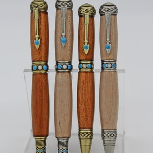 Puede incluir: Cuatro bolígrafos de madera con detalles metálicos y piedras turquesa. Los bolígrafos se muestran en un soporte de acrílico transparente. Los bolígrafos tienen diferentes acabados de madera, incluyendo un marrón rojizo y un marrón claro.