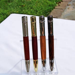Könnte beinhalten: Vier Kugelschreiber mit Holzschaft und Metallkappen, die mit einem Kruzifix-Clip versehen sind. Die Stifte stehen in einem transparenten Acrylständer. Jeder Stift hat ein einzigartiges Metallfinish in gebürstetem Silber, Gold, Chrom oder Anthrazit.