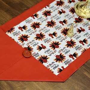 May include: A red and white table runner with a Thanksgiving turkey pattern. The pattern features turkeys with the text "Gobble Til You Wobble!" and "I Gobble 'em All!" The runner has a red border and a button on the corner.