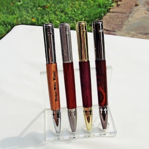 May include: Four pens with wooden barrels and metallic caps displayed on a clear acrylic stand. Each pen features a cross emblem on the clip and tip. The leftmost pen is engraved with the text Pastor Mike Nimor. The barrels vary in wood grain and color.