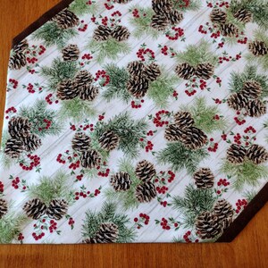 May include: A white table runner with a brown border features a repeating pattern of pine cones and holly berries on a woodgrain background.
