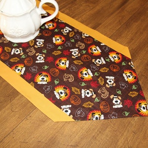May include: A brown table runner with a yellow border features a repeating pattern of cartoon dogs wearing pilgrim hats and holding turkeys. The pattern also includes pumpkins, acorns, and leaves.