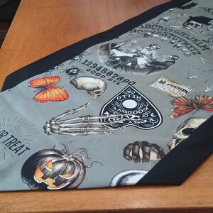 May include: A grey and black Halloween table runner with a spooky design featuring a jack-o-lantern, a skeleton hand, a butterfly, a spider web, a Ouija board, and the text "Trick or Treat".