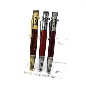Op de afbeelding: Drie bolt-action pennen met houten behuizing en metalen onderdelen. De linkerpen heeft een gouden afwerking, de middelste pen is chroom en de rechterpen heeft een gunmetal afwerking. Elke pen is voorzien van een clip en decoratieve ringen.