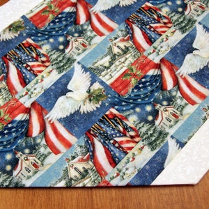 May include: A white table runner with a red, white, and blue patriotic design featuring American flags, doves, and holly. The runner has a white border with a subtle floral pattern.
