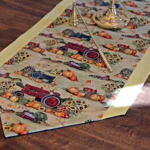 May include: A yellow and green table runner with a fall harvest theme. The runner features red and blue tractors, pumpkins, sunflowers, and baskets of apples. The runner has a yellow border.