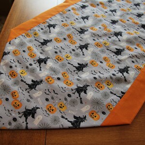 May include: A gray and orange Halloween table runner with a repeating pattern of witches, pumpkins, ghosts, and bats. The runner has a gray background with orange borders.
