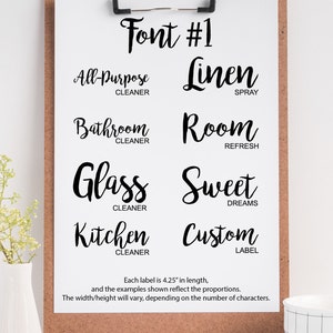 May include: A white sheet of paper with the text "Font #1" at the top. The paper is on a clipboard with a brown corkboard backing. The paper shows examples of a handwritten font with different words and phrases, such as "All-Purpose Cleaner", "Linen Spray", "Bathroom Cleaner", "Room Refresh", "Glass Cleaner", "Sweet Dreams", "Kitchen Cleaner", and "Custom Label". Each label is 4.25 inches in length.