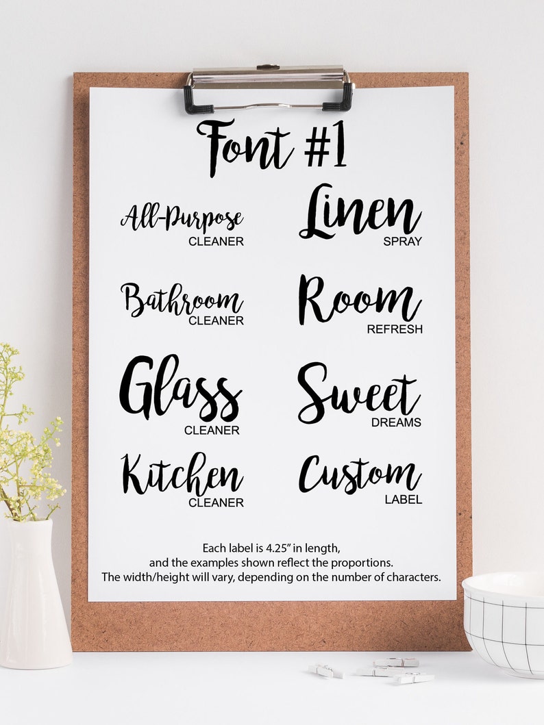 May include: A white sheet of paper with the text "Font #1" at the top. The paper is on a clipboard with a brown corkboard backing. The paper shows examples of a handwritten font with different words and phrases, such as "All-Purpose Cleaner", "Linen Spray", "Bathroom Cleaner", "Room Refresh", "Glass Cleaner", "Sweet Dreams", "Kitchen Cleaner", and "Custom Label". Each label is 4.25 inches in length.