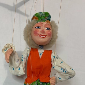 May include: A marionette puppet featuring a painted face with rosy cheeks and gray hair, wearing a white long-sleeve shirt with blue floral patterns, an orange vest, a green and orange floral skirt with yellow and white trim, and red shoes.