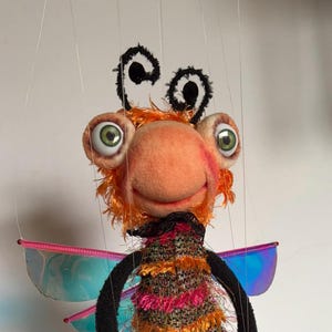 May include: A whimsical handmade puppet resembling an insect. It features a peach-colored face, large green eyes, and orange hair. The puppet wears a colorful, textured outfit, black arms and legs, and brown shoes. Iridescent wings are attached to the back.