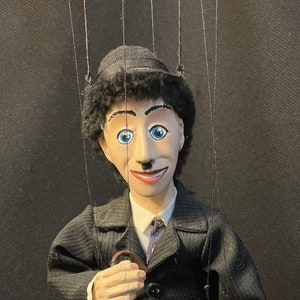 May include: A hand puppet of Charlie Chaplin, dressed in a black suit with a white shirt and a black tie. He is holding a brown cane and has a black hat on his head.