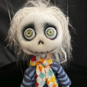 May include: A marionette puppet with a white felt skull-like head, messy white hair, and large green eyes with dark shading. The puppet wears a blue and dark blue striped long-sleeve shirt and a colorful diamond-patterned neckerchief scarf.
