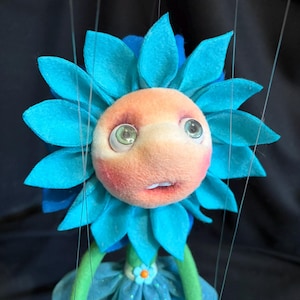 May include: A whimsical flower puppet with a turquoise petal crown and a peach-colored face. The puppet has large, expressive eyes and a small, open mouth. It wears a blue dress and has green arms and legs. The puppet is suspended by strings, suggesting it is a marionette.