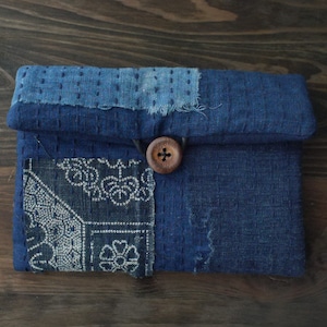 Könnte beinhalten: Eine rechteckige Tasche aus indigoblau gefärbtem Stoff mit sichtbaren weißen Steppstichen. Die Außenseite zeigt Patchwork-Elemente mit einem Blumenmuster auf der linken Seite sowie einen Holzknopfverschluss mit einer dunklen Kordelschlaufe.